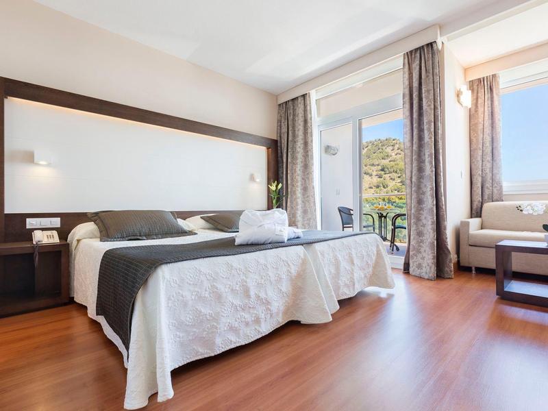 Světle hotelový pokoj s dvojlůžkem, třemi okny a dřevěnou podlahou.