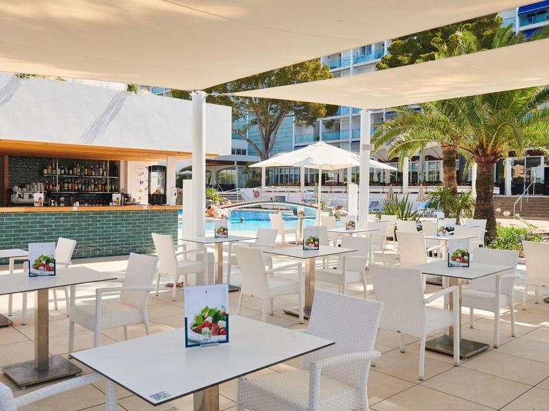 Moderní venkovní prostor hotelu se stoly a židlemi vedle bazénu pod slunečníkem.