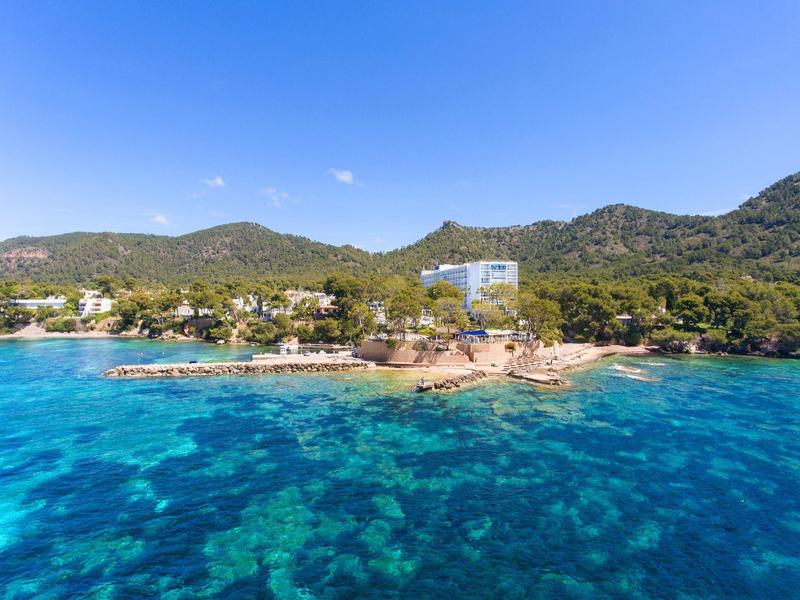 Čisté modré moře před zeleným, kopcovitým ostrovem s několika budovami na pobřeží.