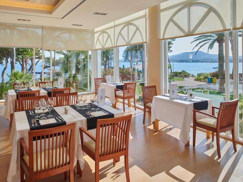 Světla restaurace s dřevěnými stoly a židlemi, velká okna s výhledem na moře a palmy.