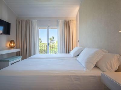 Bright hotel room with large bed, TV, desk, and balcony overlooking greenery.