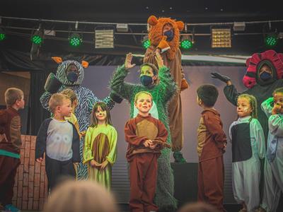 Children in animal costumes and an adult with a lion head on stage during a performance.