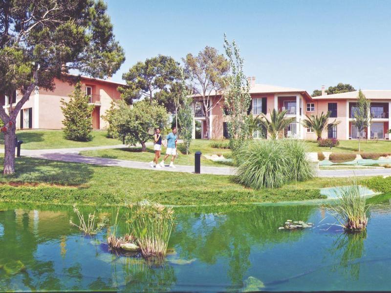 Hotel building with balcony rooms by a pond, surrounded by trees and lawn.