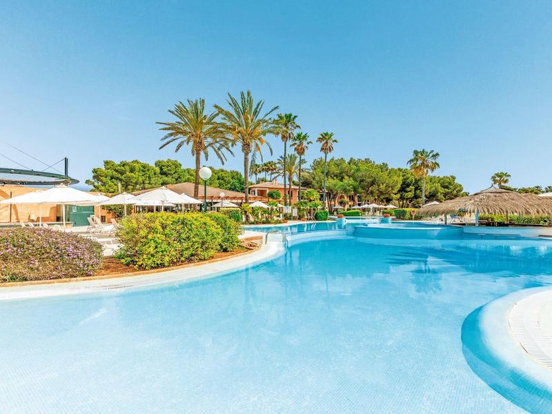 Large swimming pool surrounded by palm trees and sun umbrellas under clear blue sky