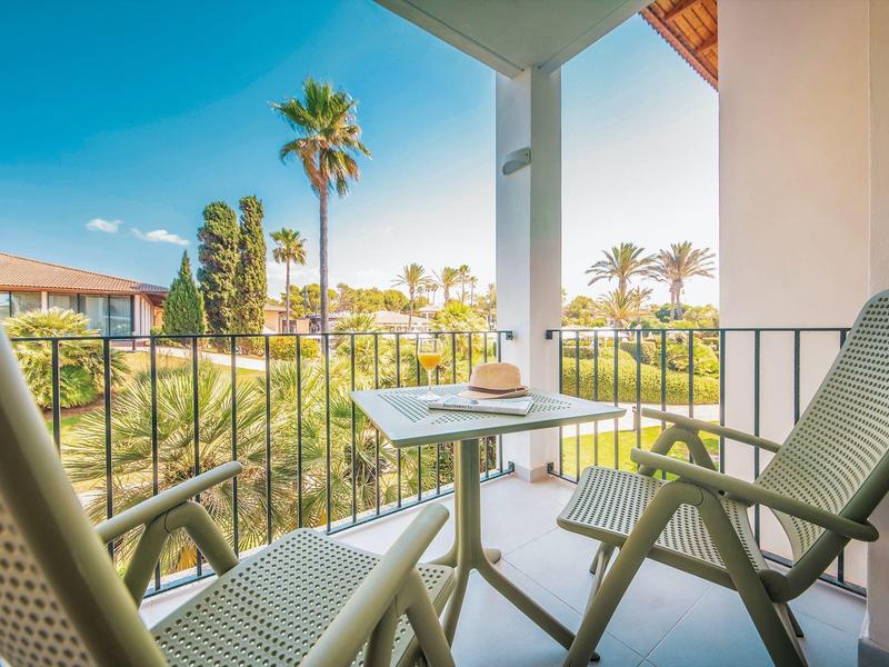 Balcony with two chairs and table, view of palm trees and hotel buildings under blue sky.