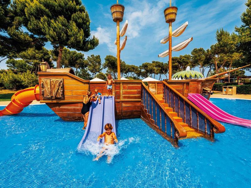 Child sliding down colorful water slides in pool with wooden ship playground.