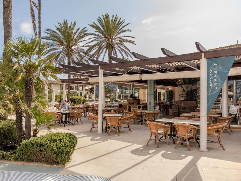 Open-air restaurant with pergola, wooden tables and chairs in a sunny outdoor area with palm trees.