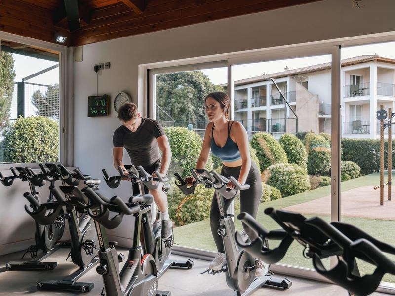 Two people exercise on indoor cycling bikes in a gym overlooking a garden area.