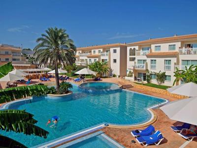 Hotel pool with lounge chairs, umbrellas, and palm trees under a clear blue sky.