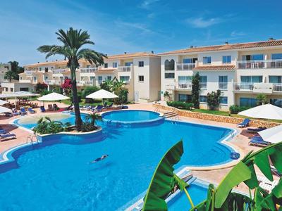 Hotel met groot gebogen zwembad, ligstoelen en witte parasols onder blauwe lucht.