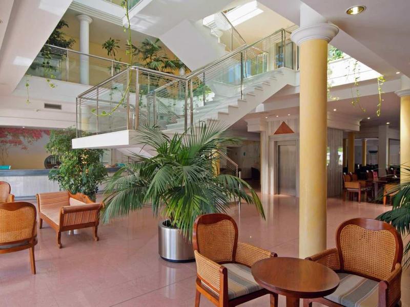 Bright hotel lobby with plants, seating areas, and staircase with glass railing.