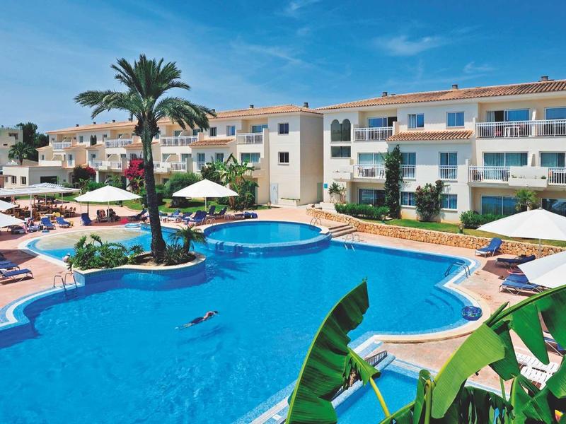 Hotel with large curved pool, sun loungers, and white umbrellas under blue sky.