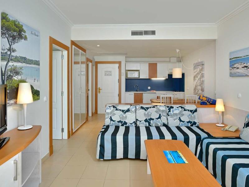 Bright hotel room with double bed, blue-striped furniture, and open kitchenette.