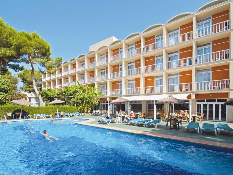 Multi-story hotel building next to large pool and sun loungers under clear sky.