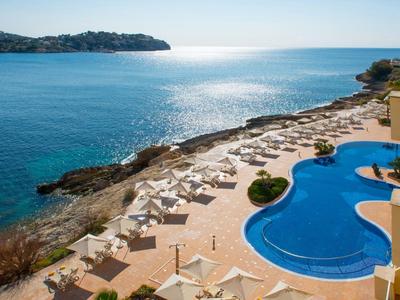 Blick auf einen Meeresstrand mit blauem Pool, weißen Liegestühlen und klarem Himmel.