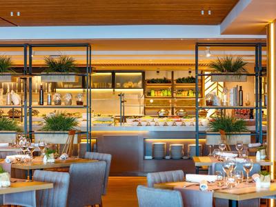 Moderner Restaurantraum mit Holzakzenten, grauen Sesseln, gedeckten Tischen und Regalen mit Pflanzen.