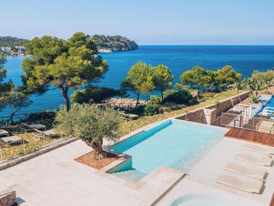 Blick auf einen Pool mit Liegen, umgeben von Bäumen und blauem Meer im Hintergrund.