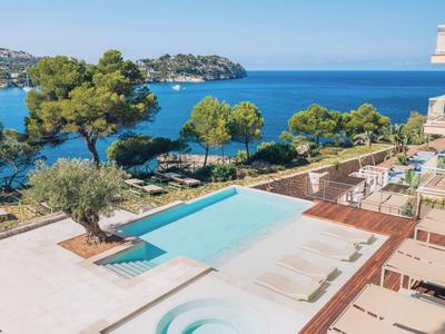 Moderner Außenpool mit Liegestühlen, umgeben von Bäumen und Blick auf das blaue Meer unter klarem Himmel.