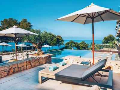 Ligbedden en parasols bij een helder zwembad met uitzicht op zee en bomen onder een heldere lucht.