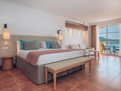 Helles Hotelzimmer mit großem Bett, Holzmöbeln, Fliesenboden und Balkon mit Meerblick.