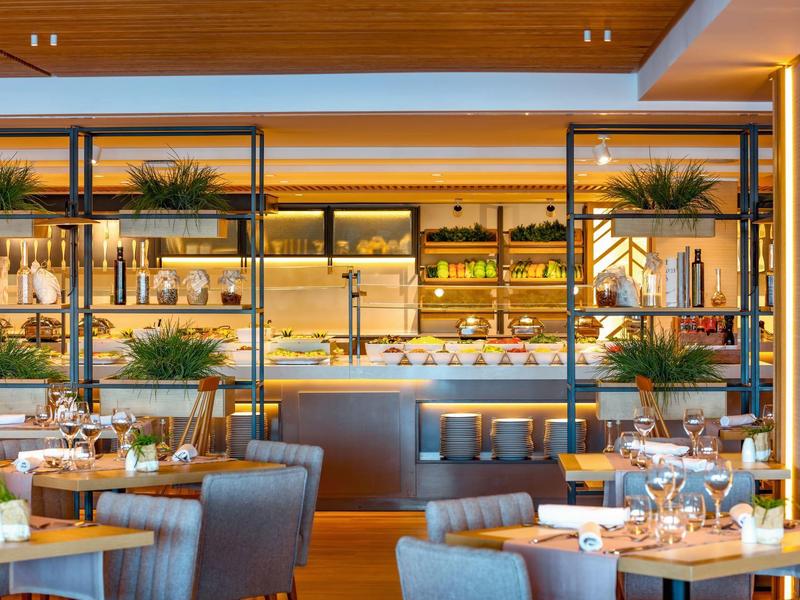 Moderner Restaurantraum mit Holzakzenten, grauen Sesseln, gedeckten Tischen und Regalen mit Pflanzen.