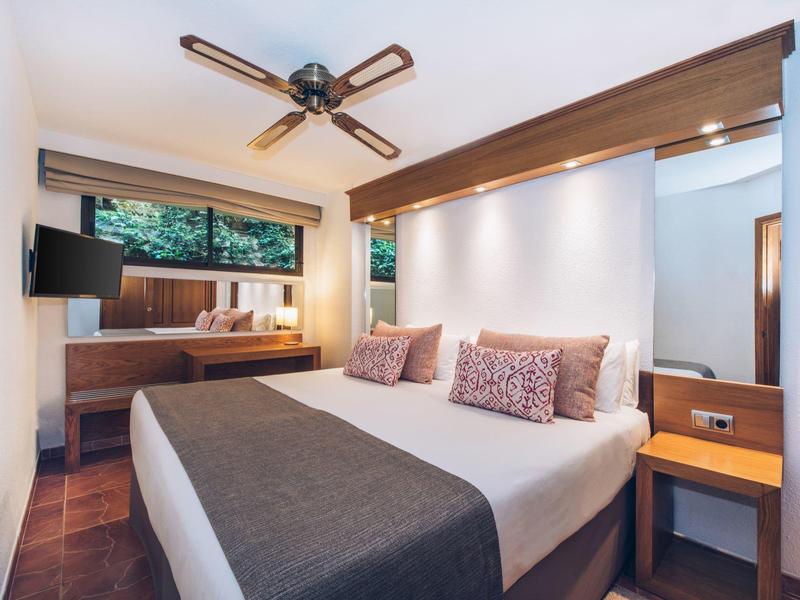 Modernes Schlafzimmer mit großem Bett, Deckenventilator, Holzmöbeln und Fenster mit Außenblick.