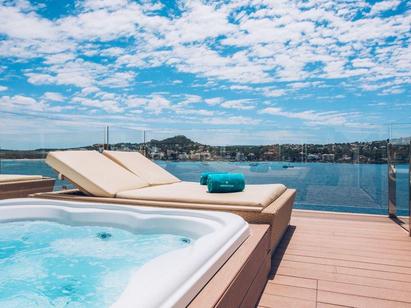 Whirlpool und Liegestühle auf Holzterrasse mit Blick auf ruhiges Meer und Hügel im Hintergrund.