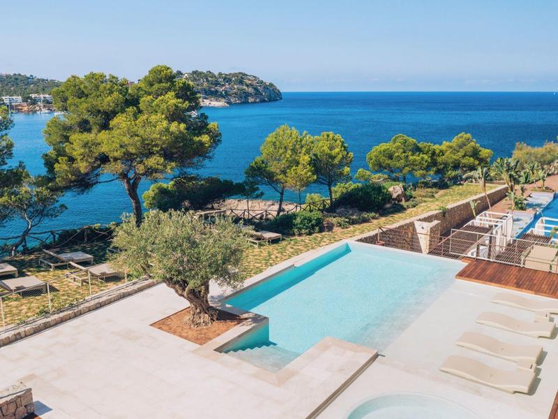 Blick auf einen Pool mit Liegen, umgeben von Bäumen und blauem Meer im Hintergrund.