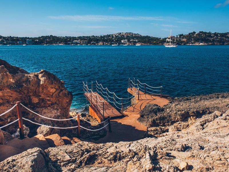 Felsige Küste mit Treppe und Geländer, blaues Meer, Inseln im Hintergrund unter klarem Himmel.
