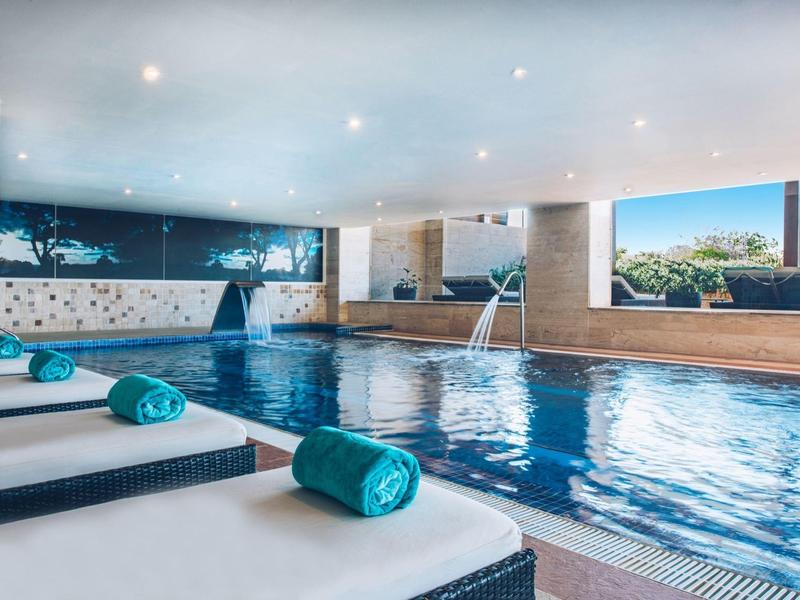 Hallenbad mit ruhigem Wasser, weiß-blauen Liegen und großen Fenstern mit Ausblick auf Bäume.
