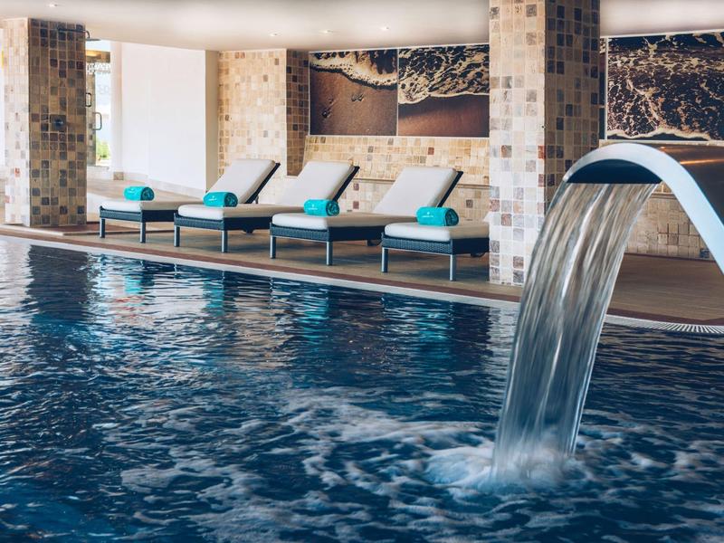 Innen-Schwimmbad mit Wasserfall und Liegen vor großen Fenstern und Mosaikfliesen.