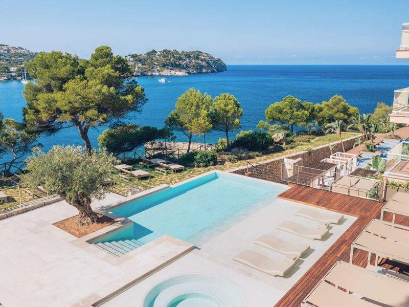 Moderner Außenpool mit Liegestühlen, umgeben von Bäumen und Blick auf das blaue Meer unter klarem Himmel.