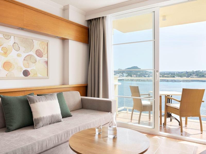 Helles Wohnzimmer mit grauer Couch, Holztisch, Kunstbild und Blick auf Balkon mit Stühlen und Meer.