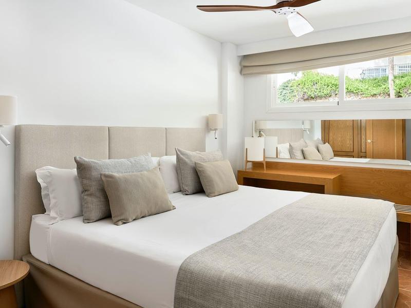 Helles Schlafzimmer mit großem Bett, grauen Kissen und Decke, Holzfensterbank mit Sitzkissen.