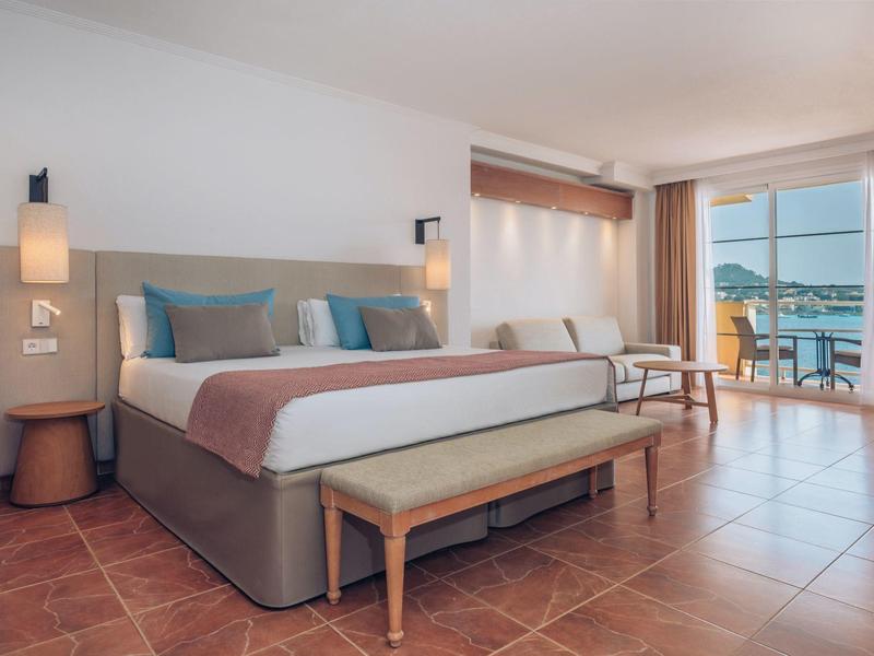 Helles Hotelzimmer mit großem Bett, Holzmöbeln, Fliesenboden und Balkon mit Meerblick.