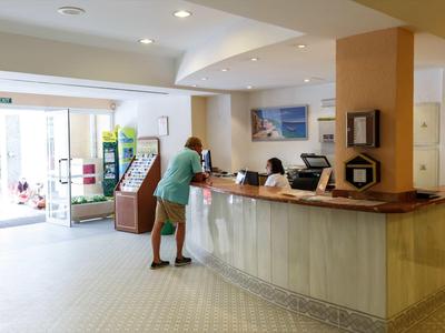 Innenraum einer Hotelrezeption mit Tresen, zwei Mitarbeitern und einem Gast in heller, freundlicher Atmosphäre.