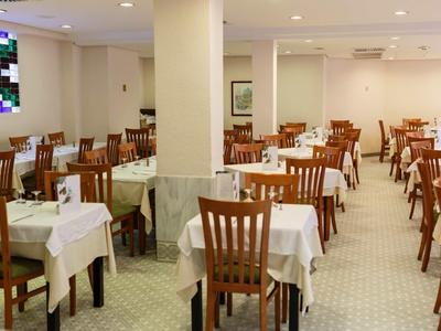 Großer, heller Speisesaal mit weißen Tischdecken und Holzstühlen, außerdem Fenster mit buntem Glas.