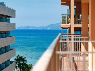 Blick vom Balkon auf das blaue Meer zwischen zwei Wohngebäuden mit Palmen und Bergen im Hintergrund.