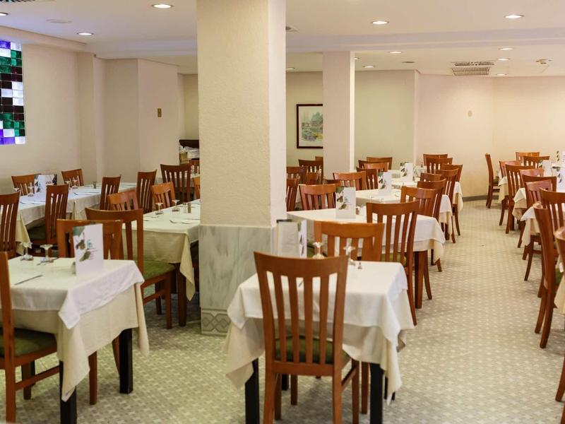 Großer, heller Speisesaal mit weißen Tischdecken und Holzstühlen, außerdem Fenster mit buntem Glas.