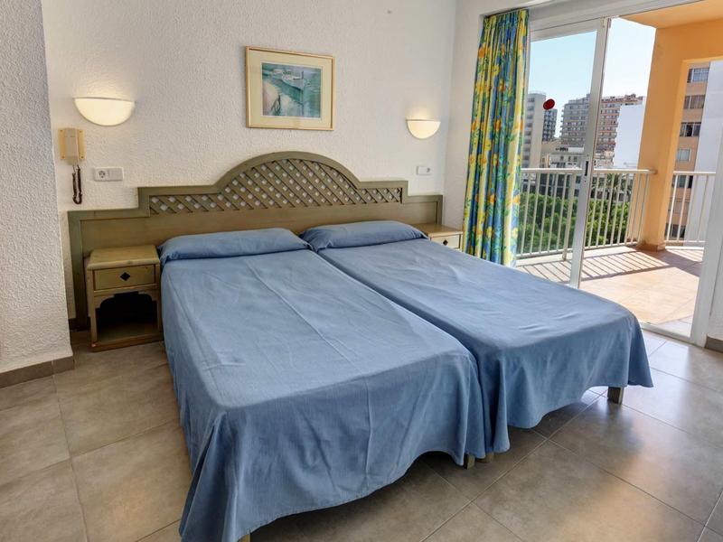 Zweibettzimmer mit blauen Bettdecken, hellen Wänden, seitlichen Nachttischen und Balkon mit Stadtblick.