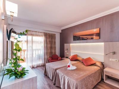 Modern hotel room with double bed, red-orange pillows, plants, and large window.
