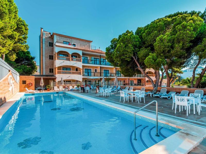 Hotel building with large outdoor pool and seating area with white chairs under trees.