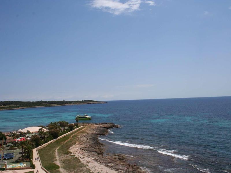 Littoral avec mer bleue claire, rochers et collines verdoyantes sous un ciel lumineux.
