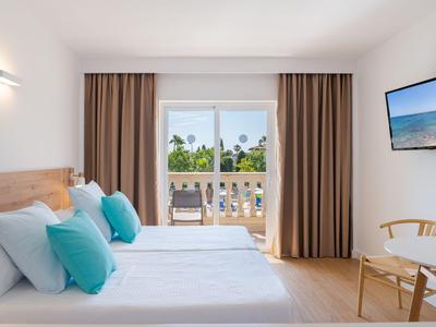 Helles Hotelzimmer mit zwei Betten, türkisfarbenen Kissen und Balkon mit Meerblick.