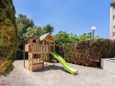 Area giochi per bambini con scivolo verde e struttura da arrampicata all'aperto accanto a siepi e un edificio.