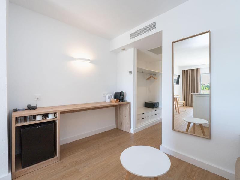 Camera d'hotel luminosa con grande specchio, mini frigorifero e armadio aperto.