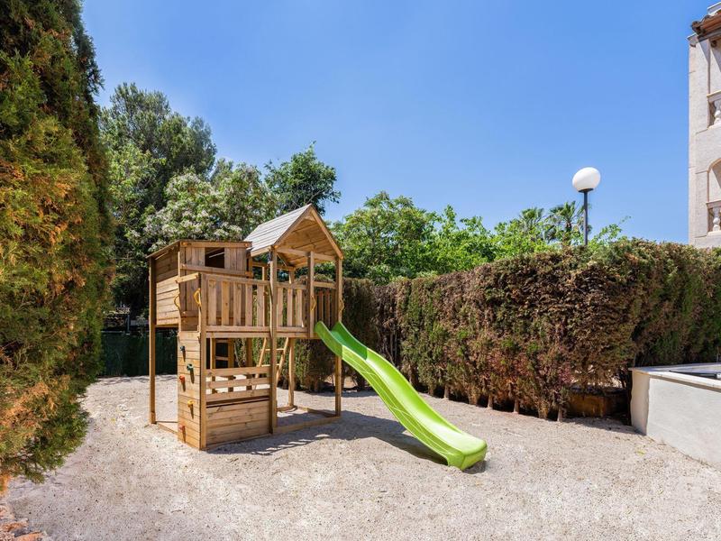 Area giochi per bambini con scivolo verde e struttura da arrampicata all'aperto accanto a siepi e un edificio.