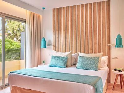 Helles Hotelzimmer mit Doppelbett, türkisfarbenen Akzenten und Balkon mit Pflanzenblick.