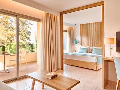 Helles Hotelzimmer mit großem Bett, Balkon und Tisch mit Stuhl, warmes Holzakzent-Design.