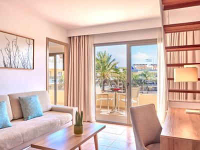 Helles Hotelzimmer mit Sofa, Holztisch, Schreibtisch und Blick auf Terrasse mit Palmen.